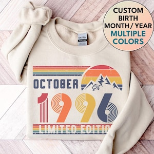 May include: A cream-coloured crewneck sweatshirt featuring a graphic print with the text OCTOBER 1996 LIMITED EDITION. The design includes a retro-style sunset over mountain peaks in shades of red, orange, yellow, and blue, with striped accents.
