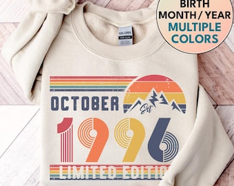 May include: A cream-colored crewneck sweatshirt featuring a graphic print with the text OCTOBER 1996 LIMITED EDITION. The design includes a retro-style sunset over mountain peaks in shades of red, orange, yellow, and blue, with striped accents.