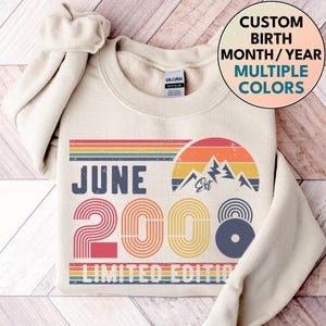 Peut inclure: Sweat-shirt crème avec un design rétro. Le motif comprend le texte "JUNE 2000 LIMITED EDITION" avec un arc-en-ciel, un graphique de montagne et un motif circulaire. Le sweat-shirt porte également le texte "CUSTOM BIRTH MONTH/YEAR MULTIPLE COLORS".