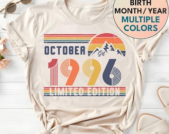 May include: A cream-colored t-shirt featuring a graphic with the text OCTOBER 1996 LIMITED EDITION. The design includes a retro-style sunset, mountain peaks, and horizontal stripes in red, orange, yellow, and blue. The text CUSTOM BIRTH MONTH/YEAR MULTIPLE COLORS is also visible.