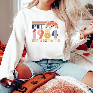 May include: White long-sleeve sweatshirt with the text "APRIL 1984 LIMITED EDITION" in a retro design. The graphic includes a mountain range, a sunset, and the year 1984 in a colorful, vintage style. The sweatshirt is a casual, everyday garment.