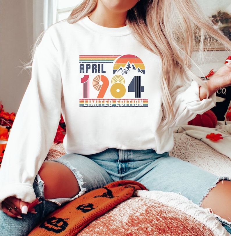 May include: White long-sleeve sweatshirt with the text "APRIL 1984 LIMITED EDITION" in a retro design. The graphic includes a mountain range, a sunset, and the year 1984 in a colorful, vintage style. The sweatshirt is a casual, everyday garment.