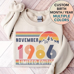 May include: A cream-colored crewneck sweatshirt featuring a graphic print with the text NOVEMBER 1986 LIMITED EDITION. The design includes a retro-style sunset, mountain silhouettes, and horizontal stripes in shades of red, orange, yellow, and blue.