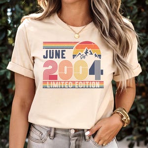 以下が含まれることがあります： JUNE 2004 LIMITED EDITIONというテキストがプリントされたクリーム色のTシャツ。レトロなスタイルの夕日、山のシルエット、赤、オレンジ、黄色、青の横縞模様がデザインされています。