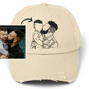 Puede incluir: Una gorra de béisbol desgastada de color beige con un dibujo de línea negra de un padre y un hijo abrazándose. El dibujo está en la parte delantera de la gorra.