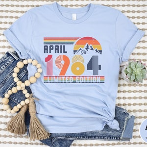 May include: A light blue t-shirt featuring a graphic print with the text "APRIL 1984 LIMITED EDITION" and "EST". The design includes a retro-style sunset, mountain silhouettes, and horizontal stripes in shades of red, orange, yellow, and dark blue.
