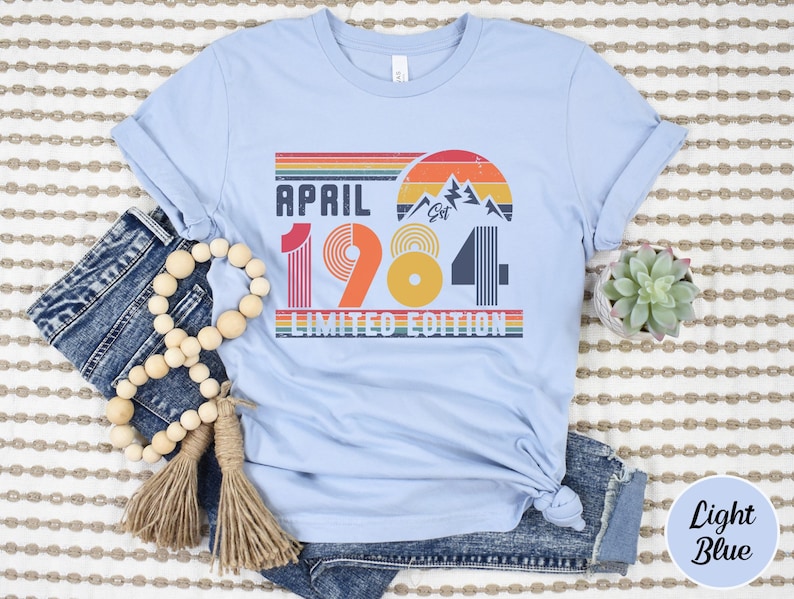 May include: A light blue t-shirt featuring a graphic print with the text "APRIL 1984 LIMITED EDITION" and "EST". The design includes a retro-style sunset, mountain silhouettes, and horizontal stripes in shades of red, orange, yellow, and dark blue.