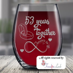 May include: A clear wine glass with a red liquid inside. The glass is engraved with the text "53 years together forever to go" and two intertwined rings. There are two small hearts on the glass. The text "All rights reserved by Freedom Look" is printed on a white label at the bottom of the glass.