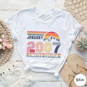 Op de afbeelding: Een wit t-shirt met een grafische print met de tekst JANUARY 2007 LIMITED EDITION. Het ontwerp bevat een retro zonsondergang, bergsilhouetten en horizontale strepen in de kleuren rood, oranje, geel, groen en blauw.