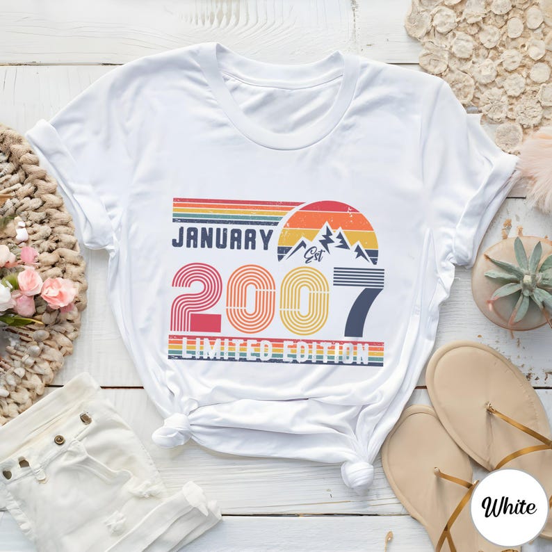 Op de afbeelding: Een wit t-shirt met een grafische print met de tekst JANUARY 2007 LIMITED EDITION. Het ontwerp bevat een retro zonsondergang, bergsilhouetten en horizontale strepen in de kleuren rood, oranje, geel, groen en blauw.
