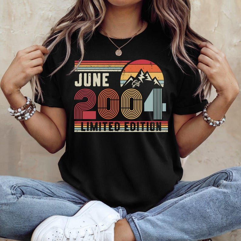 以下が含まれることがあります： 夕日、山々、そして「JUNE 2004 LIMITED EDITION」という文字が描かれたレトロなスタイルのグラフィックが特徴の黒い半袖Tシャツ。デザインには、オレンジ、黄色、赤、青の色合いの水平ストライプが使用されており、ヴィンテージ感のある仕上がりです。