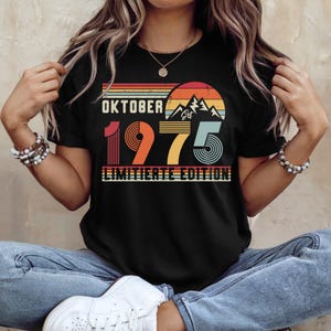 May include: A black short-sleeved t-shirt featuring a graphic design with the text "OKTOBER 1975 LIMITIERTE EDITION" and "Est". The design includes a retro-style sunset, mountain silhouettes, and horizontal stripes in shades of orange, yellow, and teal.