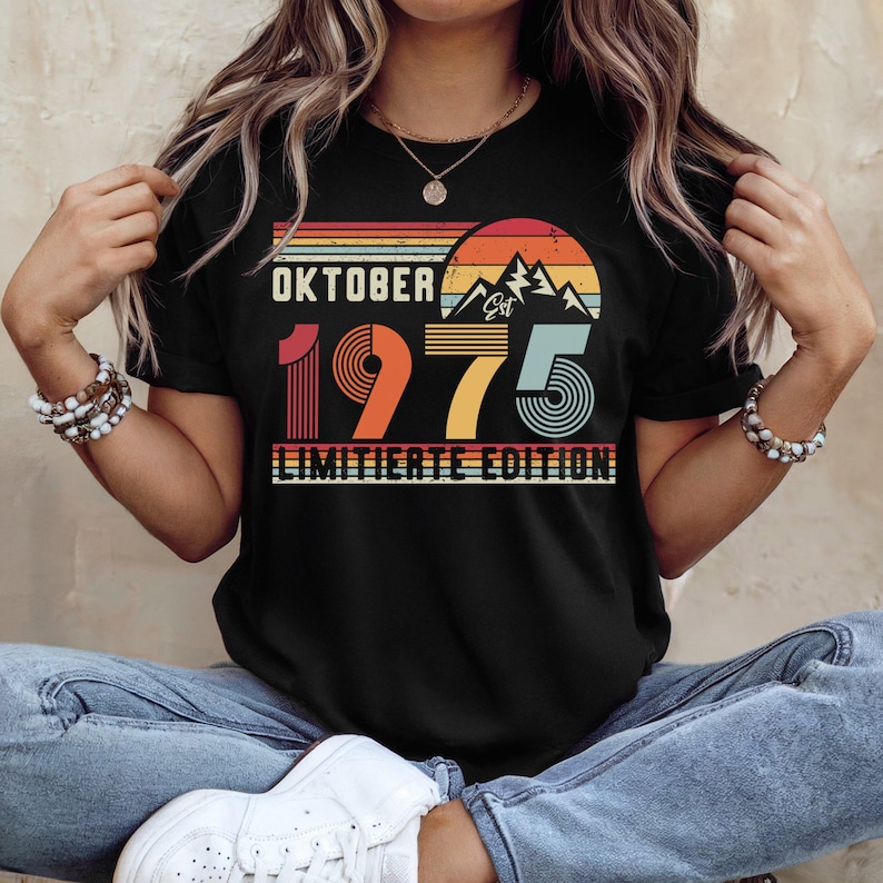 May include: A black short-sleeved t-shirt featuring a graphic design with the text "OKTOBER 1975 LIMITIERTE EDITION" and "Est". The design includes a retro-style sunset, mountain silhouettes, and horizontal stripes in shades of orange, yellow, and teal.