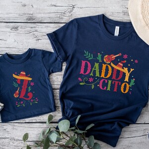 May include: Two navy blue t-shirts with a festive Mexican theme. The larger shirt says "Daddy Cito" with a sombrero, guitar, and cactus design. The smaller shirt has a sombrero and guitar design with the letter "L".