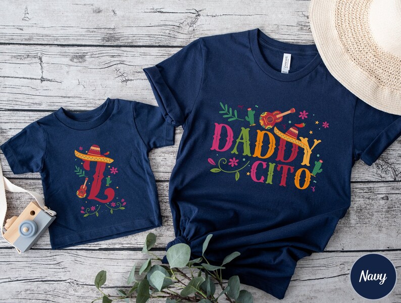 May include: Two navy blue t-shirts with a festive Mexican theme. The larger shirt says "Daddy Cito" with a sombrero, guitar, and cactus design. The smaller shirt has a sombrero and guitar design with the letter "L".