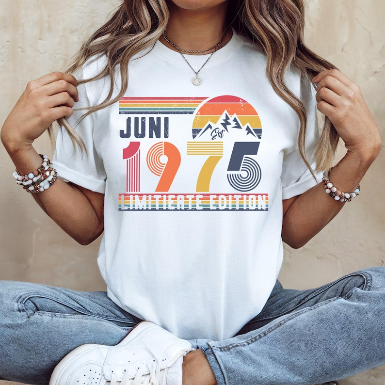 May include: A white t-shirt featuring a retro-style graphic with the text JUNI 1975 LIMITIERTE EDITION. The design includes a sunset, mountain peaks, and horizontal stripes in shades of red, orange, yellow, and blue, with the word Est printed above 1975.