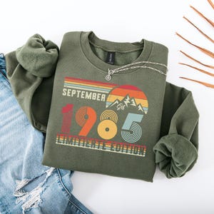 May include: An olive green crewneck sweatshirt featuring a graphic print with the text SEPTEMBER 1985 LIMITIERTE EDITION. The design includes a retro-style sunset, mountain silhouettes, and concentric circle patterns in shades of orange, yellow, and blue.