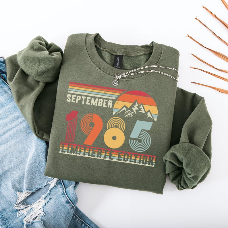 May include: An olive green crewneck sweatshirt featuring a graphic print with the text SEPTEMBER 1985 LIMITIERTE EDITION. The design includes a retro-style sunset, mountain silhouettes, and concentric circle patterns in shades of orange, yellow, and blue.
