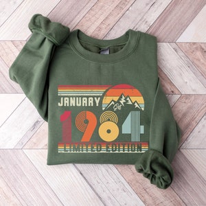 May include: A dark green sweatshirt with the text "January 1984 Limited Edition" in a retro design. The design includes a mountain range, a sunset, and a series of colorful stripes. The sweatshirt has cuffed sleeves.