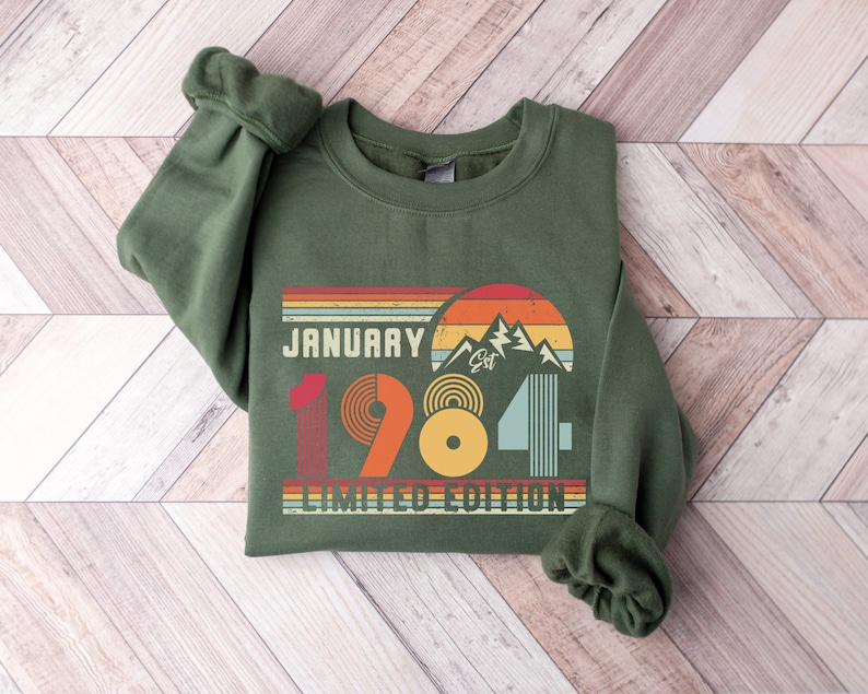 May include: A dark green sweatshirt with the text "January 1984 Limited Edition" in a retro design. The design includes a mountain range, a sunset, and a series of colorful stripes. The sweatshirt has cuffed sleeves.