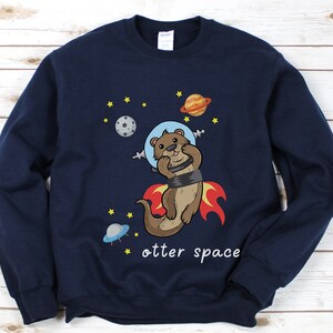 May include: A navy blue sweatshirt featuring a cartoon otter wearing a space helmet and riding a rocket. The text "otter space" is printed below the otter.