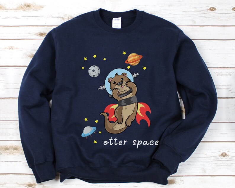 May include: A navy blue sweatshirt featuring a cartoon otter wearing a space helmet and riding a rocket. The text "otter space" is printed below the otter.