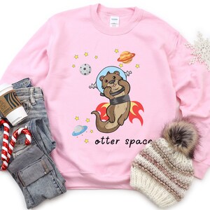 May include: Pink sweatshirt featuring a cartoon otter wearing a space helmet and flying through space. The text "otter space" is printed below the otter.