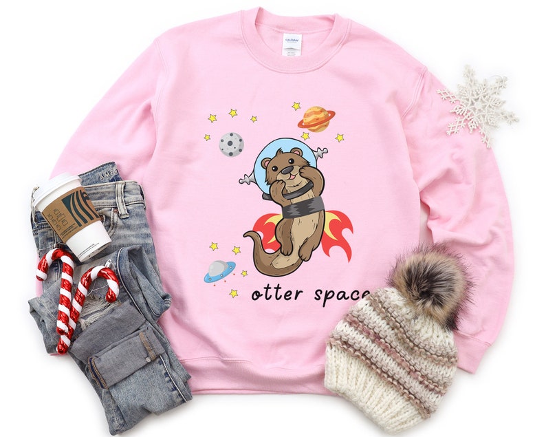 May include: Pink sweatshirt featuring a cartoon otter wearing a space helmet and flying through space. The text "otter space" is printed below the otter.