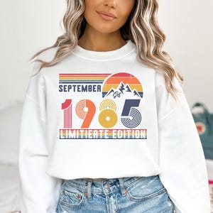 May include: A white crewneck sweatshirt featuring a graphic print with the text SEPTEMBER 1985 LIMITIERTE EDITION. The design includes a retro sunset, mountain silhouette, and horizontal stripes in shades of red, orange, yellow, and blue.