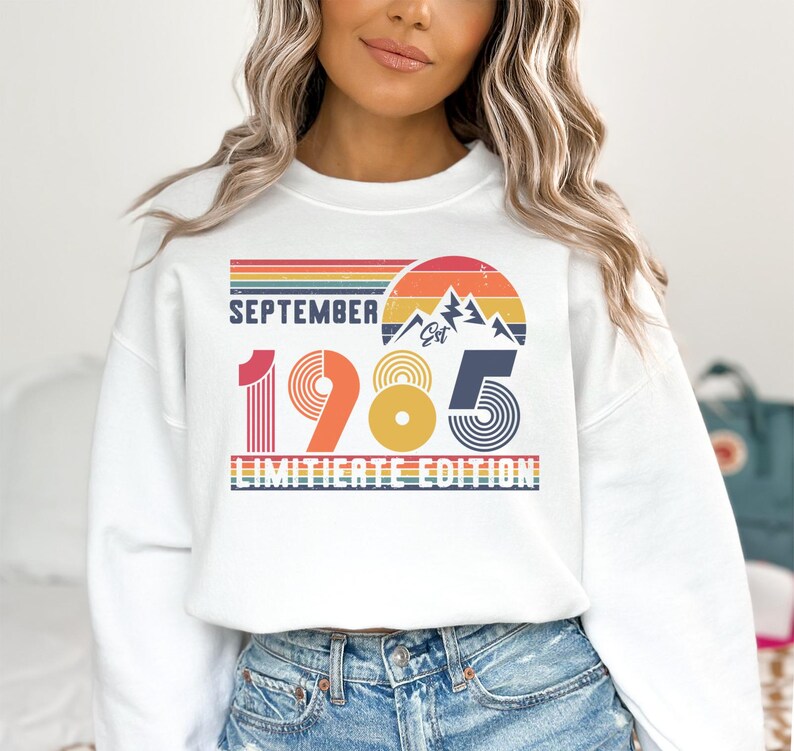 May include: A white crewneck sweatshirt featuring a graphic print with the text SEPTEMBER 1985 LIMITIERTE EDITION. The design includes a retro sunset, mountain silhouette, and horizontal stripes in shades of red, orange, yellow, and blue.