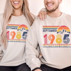 May include: A beige crewneck sweatshirt featuring a retro-style graphic with the text "SEPTEMBER 1985 LIMITED EDITION". The design includes a sunset over mountains, horizontal stripes, and stylized numbers in shades of red, orange, yellow, and blue.
