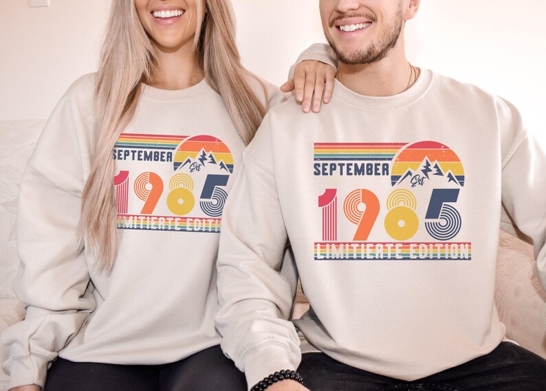 May include: A beige crewneck sweatshirt featuring a retro-style graphic with the text "SEPTEMBER 1985 LIMITED EDITION". The design includes a sunset over mountains, horizontal stripes, and stylized numbers in shades of red, orange, yellow, and blue.