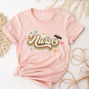 Op de afbeelding: Lichtroze t-shirt met het woord "Nurse" in een retro lettertype, versierd met een spook, vleermuis en bloemen. Het shirt is gemaakt van zacht materiaal en heeft opgerolde mouwen.