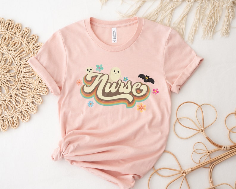 Op de afbeelding: Lichtroze t-shirt met het woord "Nurse" in een retro lettertype, versierd met een spook, vleermuis en bloemen. Het shirt is gemaakt van zacht materiaal en heeft opgerolde mouwen.