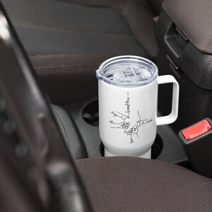 May include: A white travel mug with a handle and a clear lid, resting in a car's cup holder. The mug features a black line drawing of hands holding each other, with names and the words "Grand-P&egrave;re" written on it.