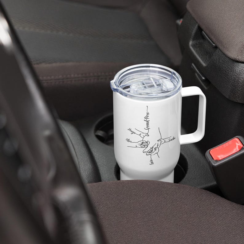 May include: A white travel mug with a handle and a clear lid, resting in a car's cup holder. The mug features a black line drawing of hands holding each other, with names and the words "Grand-P&egrave;re" written on it.