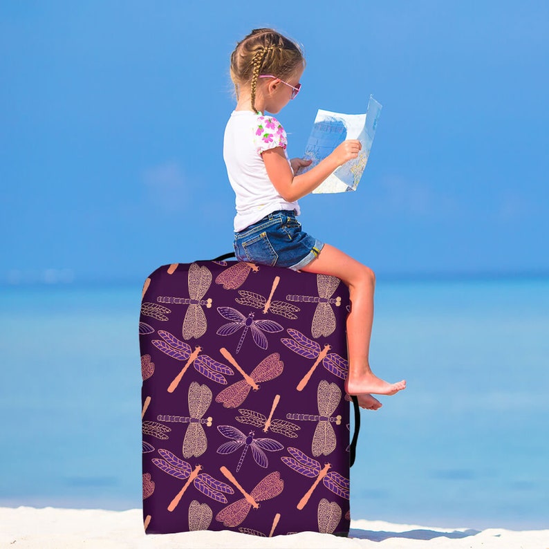 May include: Purple luggage cover with a dragonfly pattern. The dragonfly design is in shades of pink and gold. A young person is sitting on the luggage, looking at a map.