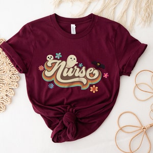 Op de afbeelding: Bordeauxrood T-shirt met opgerolde mouwen. Het shirt toont het woord "Nurse" in een retro lettertype, vergezeld van spook-, schedel-, vleermuis- en bloemenafbeeldingen. Het T-shirt is aan de onderkant geknoopt.