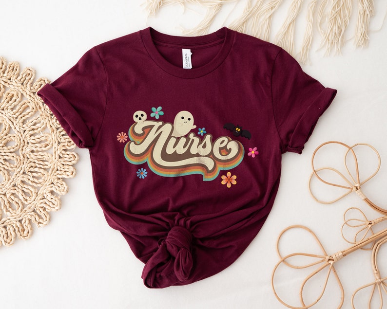 Op de afbeelding: Bordeauxrood T-shirt met opgerolde mouwen. Het shirt toont het woord "Nurse" in een retro lettertype, vergezeld van spook-, schedel-, vleermuis- en bloemenafbeeldingen. Het T-shirt is aan de onderkant geknoopt.