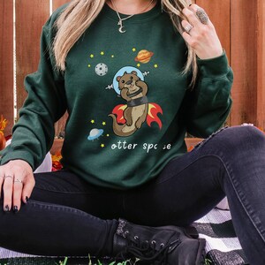 May include: A dark green sweatshirt featuring a cartoon otter wearing a space helmet and flying through space. The text "otter space" is printed below the otter.