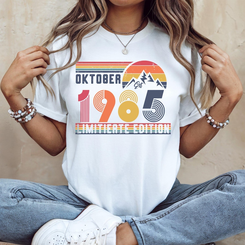 May include: A white short-sleeve t-shirt featuring a retro-style graphic with the text OKTOBER 1985 LIMITIETE EDITION. The design includes a sunset, mountain silhouettes, and striped lines in shades of orange, yellow, blue, and red.
