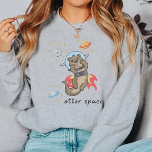 May include: Grey sweatshirt featuring a cartoon otter wearing a space helmet and flying through space. The text "otter space" is printed below the otter.
