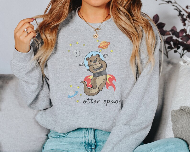 May include: Grey sweatshirt featuring a cartoon otter wearing a space helmet and flying through space. The text "otter space" is printed below the otter.
