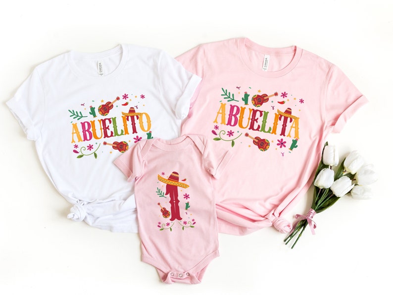 May include: Three pink and white t-shirts with a colourful floral design and the words "Abuelito" and "Abuelita" printed on them. The shirts are for a family of three.