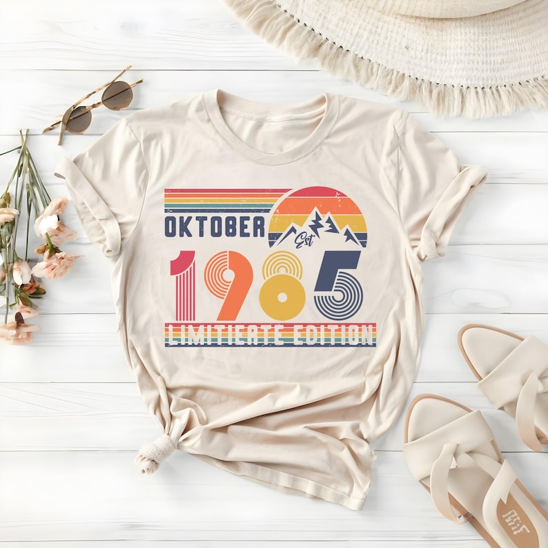 May include: A beige short-sleeve t-shirt featuring a graphic print with the text "OKTOBER 1985 LIMITIATE EDITION" and "EST". The design includes a retro-style sunset, mountain silhouettes, and horizontal stripes in shades of red, orange, and blue.