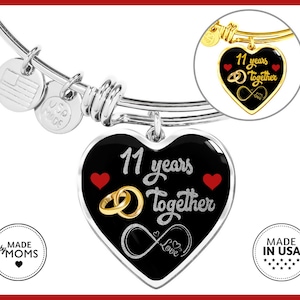 May include: A silver bangle bracelet with a black heart charm that says "11 years together" with a gold infinity symbol and the word "love". The charm also has two red hearts and two gold rings. The bracelet has a silver tag that says "Made by Moms" and a silver tag with a US flag.