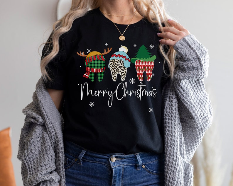 Op de afbeelding: Zwart T-shirt met een "Merry Christmas" afbeelding. Het ontwerp toont drie tanden gekleed in feestelijke vakantiekleding: een rendier, een luipaardprint hoed en een kerstboom. Sneeuwvlokken zijn verspreid rond de tekst.