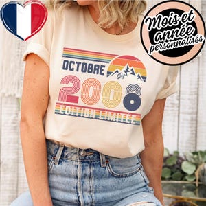 Peut inclure: Un t-shirt couleur crème arborant un graphisme rétro avec le texte OCTOBRE 2008 ÉDITION LIMITÉE. Le motif comprend un coucher de soleil, des sommets montagneux et des rayures horizontales dans des tons orange, jaune, bleu et vert, avec le mot Est.