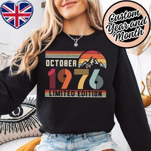 Può includere: Una felpa nera a maniche lunghe con una grafica in stile retrò raffigurante un tramonto, montagne e il testo "OCTOBER 1976 LIMITED EDITION". Il design presenta strisce orizzontali nei toni dell'arancione, del giallo e del verde acqua con estetica vintage.