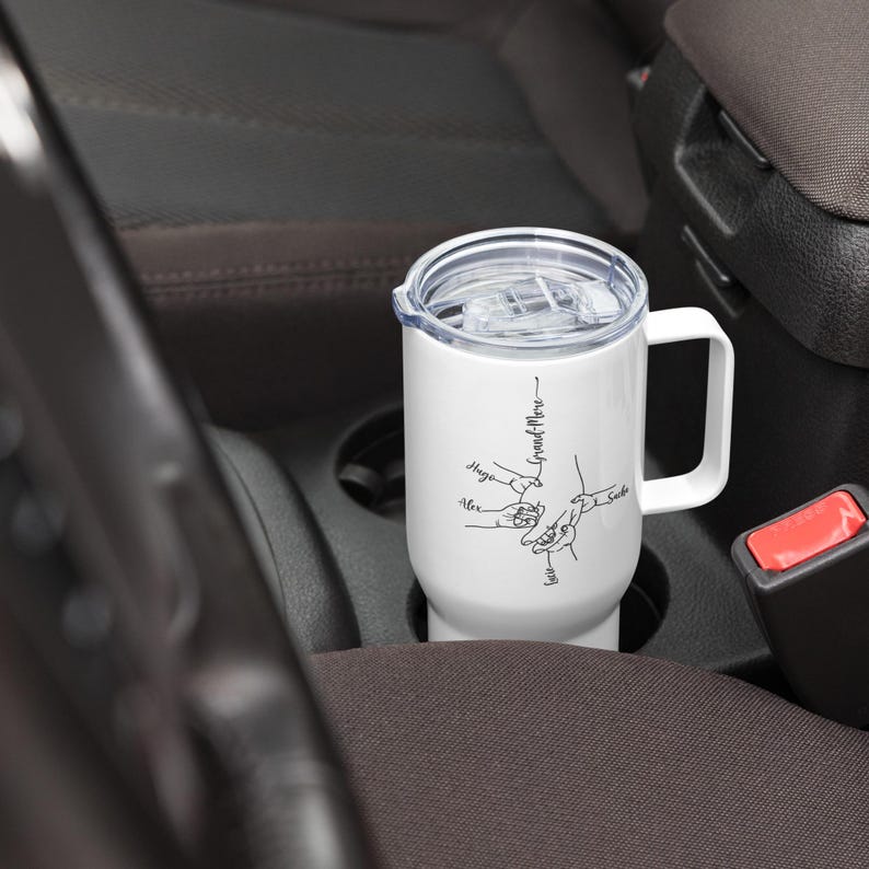 May include: White travel mug with a handle and a clear lid, resting in a car's cup holder. The mug features a black line drawing of hands and the words "Grand-Mere" and names like "Alex" and "Sage".